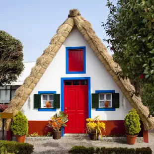 Small rural house with a triangular thatched roof, a red front door and small windows with shutters.