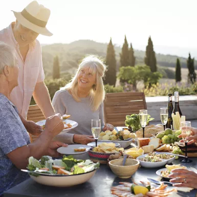 A group of mature friends are sitting around an outdoor dining table in Tuscany, Italy