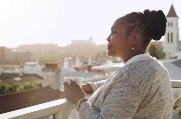 Senior woman standing alone on her balcony, contemplating and enjoying a cup of coffee in the sun