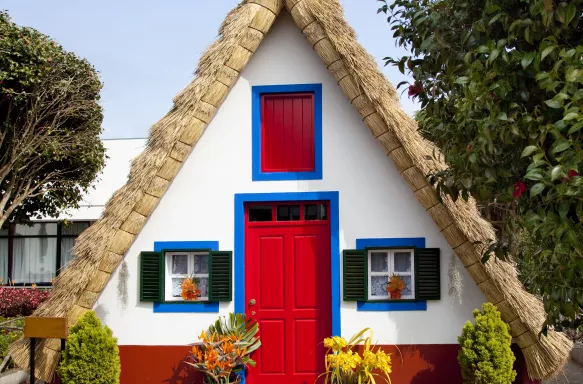 Small rural house with a triangular thatched roof, a red front door and small windows with shutters.