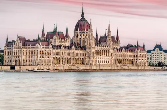 Wide angle shot of the Hungarian Parliament on the banks of the Danube River in Budapest, against a pink tinted sky.