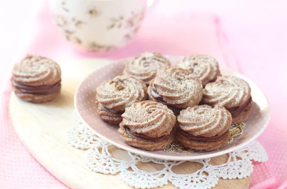 Viennese Sable Cookies with Chocolate Filling on a wooden cutting board and a cup of tea, on a light pink background.