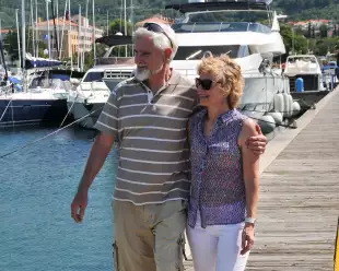 A male and female couple stood on the end of a jetty holding one another round the waist and arm with the sun on their faces.