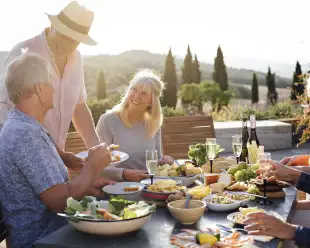 A group of mature friends are sitting around an outdoor dining table in Tuscany, Italy