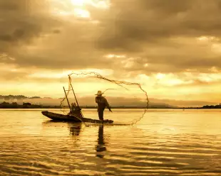 A fisherman in a boat throwing a net on the lake during sunset