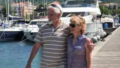 A male and female couple stood on the end of a jetty holding one another round the waist and arm with the sun on their faces.