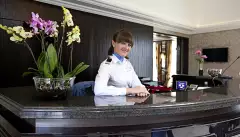 Internal shot of woman in uniform politely smiling behind a glossy, black reception desk inside the William Shakespeare cruise ship