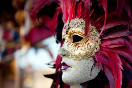 Red Venetian Carnival mask, the mask is the main focus of the picture.The mask has pink, purple and red feathers, a golden mask, a porcelain face with texture and cracks and red lips. Other blurred masks are in the background of the picture